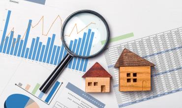 New Report on the US Housing Market Finds Record Numbers Across Three Key Metrics A magnifying glass and small wooden houses laying on paperwork with graphs on it
