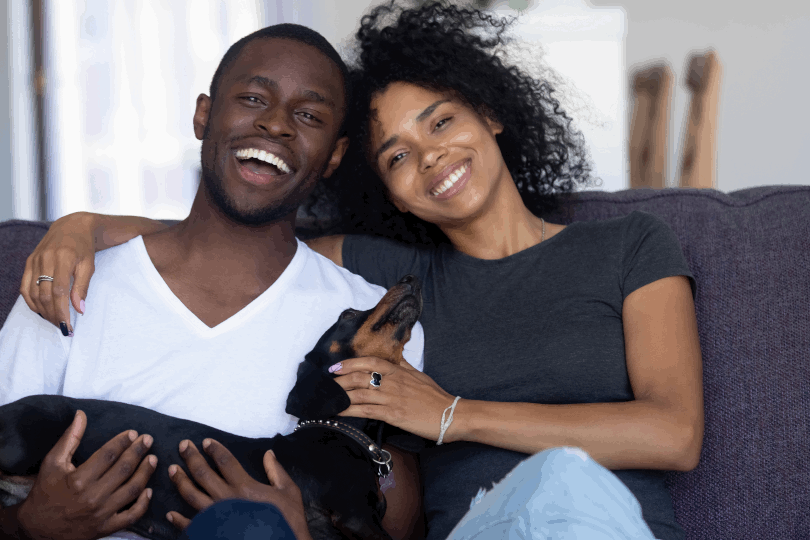 A young happy couple sitting on a couch smiling and embracing a dog